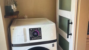 Un lave-linge compact et blanc, doté d’un panneau de commande numérique, installé dans un petit espace clos avec une porte en bois et une étagère sur laquelle se trouve une plante.