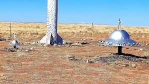 A desert landscape with a tall, cylindrical structure with a pointed top, a dome-shaped structure with an alien figure on top, and a barbed wire fence in the background. - Alien Acres UFO campground,  car, truck, RV, tent off-grid camping (Sunvalley)