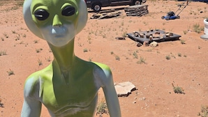 A green alien figure in a desert setting with a pickup truck and solar panels in the background. - Alien Acres UFO campground,  car, truck, RV, tent off-grid camping (Sunvalley)