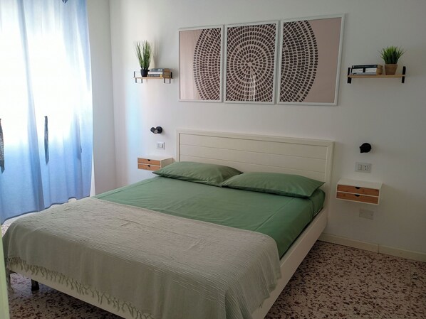 A neatly arranged bedroom with a bed, a bedside table, a wall-mounted shelf with a plant, and a wall art piece.