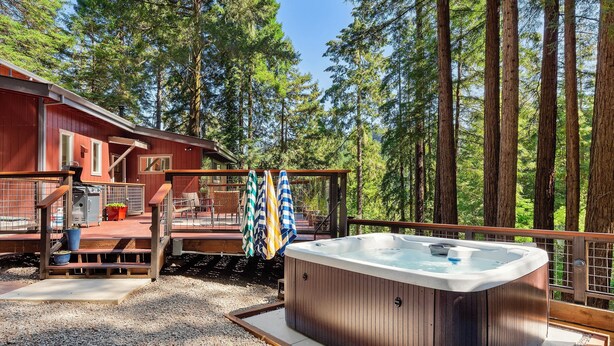 Een houten terras met een jacuzzi, omringd door hoge bomen, en een rood gebouw op de achtergrond.