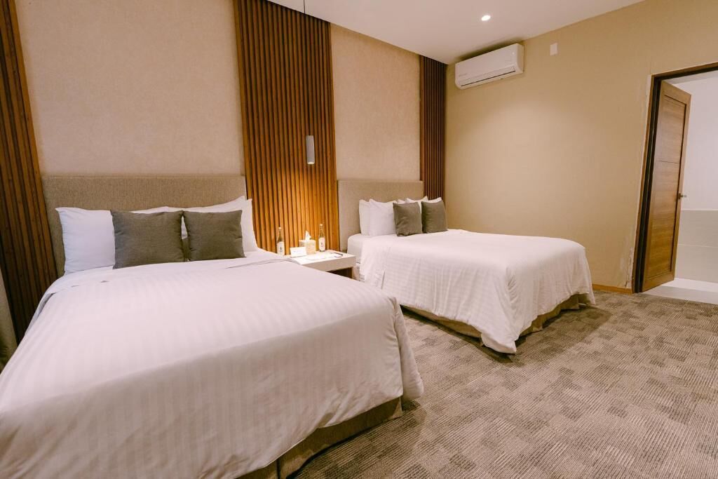 A hotel room with two beds, a wooden panel wall, an air conditioning unit, and a carpeted floor.