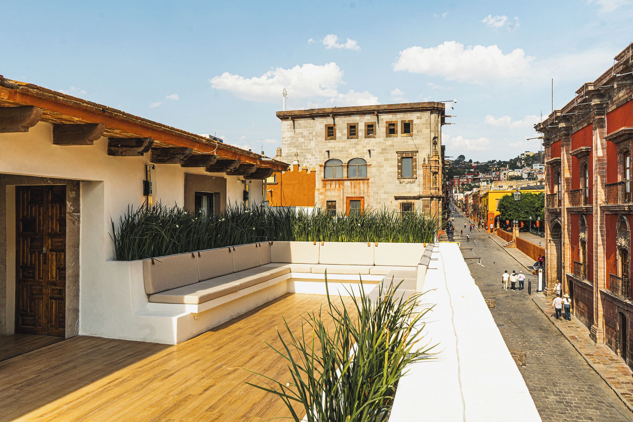 A rooftop terrace with a wooden floor, a white planter with plants, and a view of the street below.