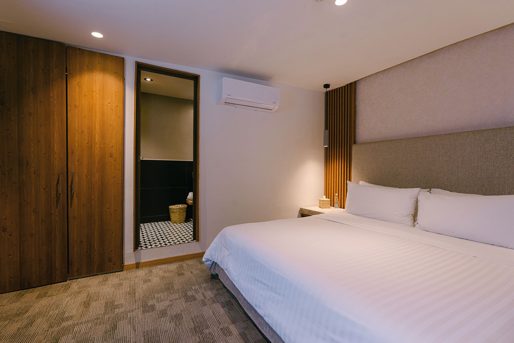 Habitación de hotel con una cama grande, un armario de madera, una mesita con una botella, aire acondicionado de pared y piso con estampado.
