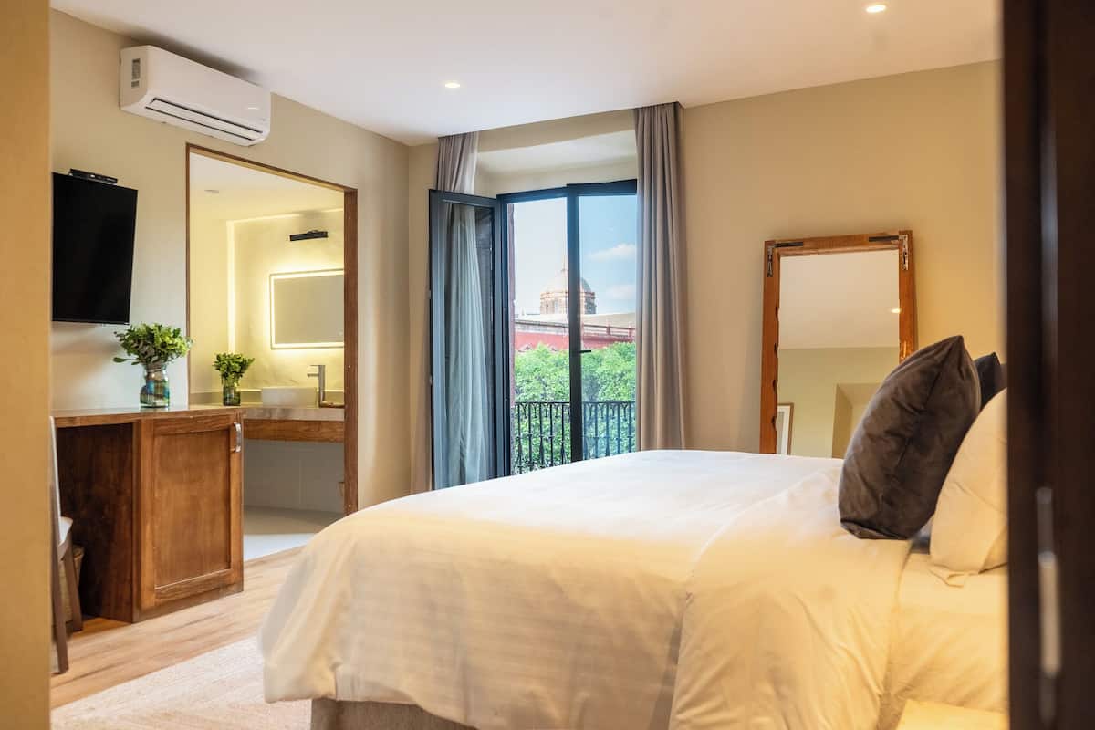A hotel bedroom with a large bed, a wooden desk, a mirror, and a view of a building through the window.