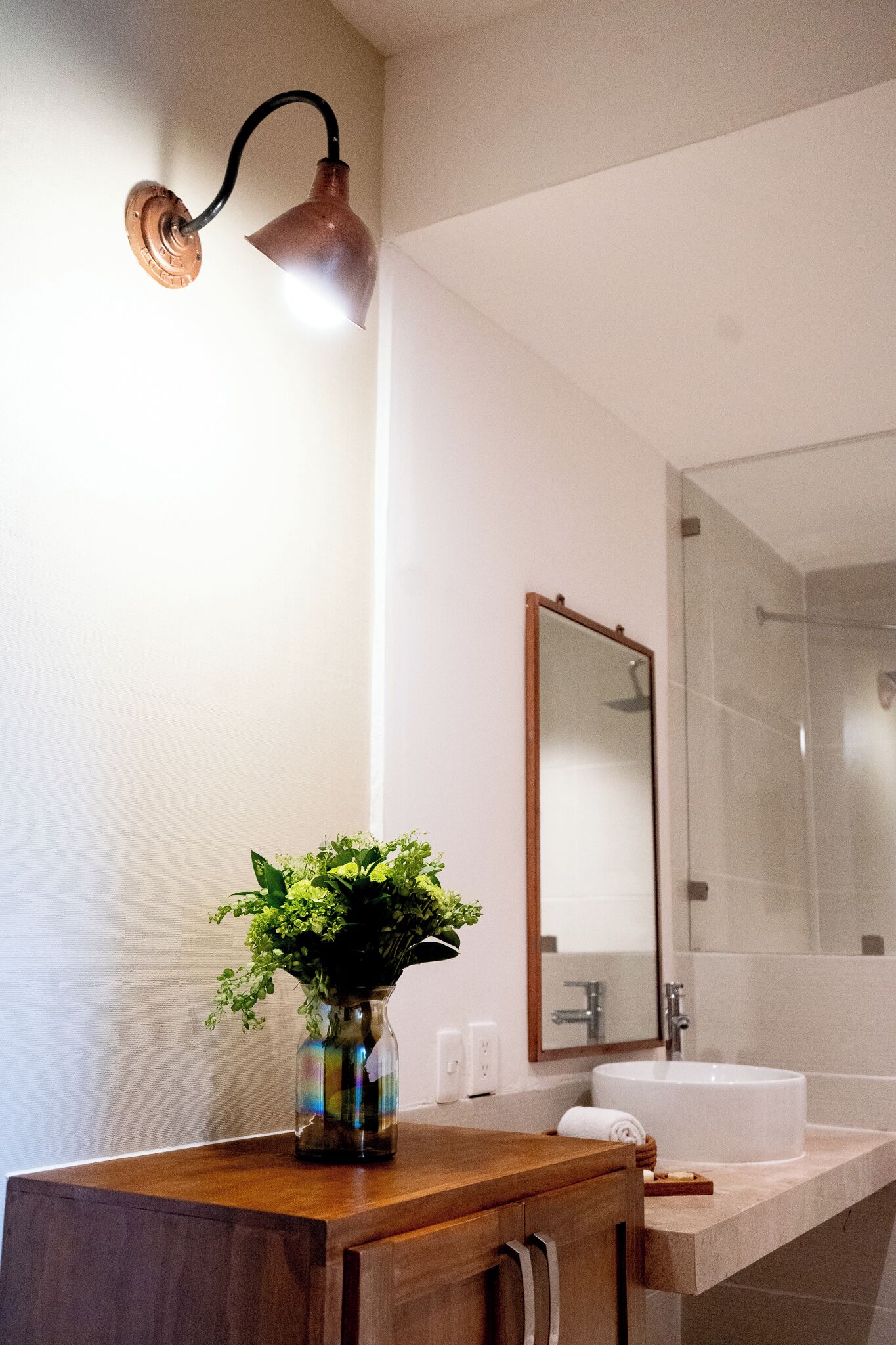 A bathroom with a wooden vanity, a vase with green plants, a wall-mounted light, a large mirror, and a white sink.