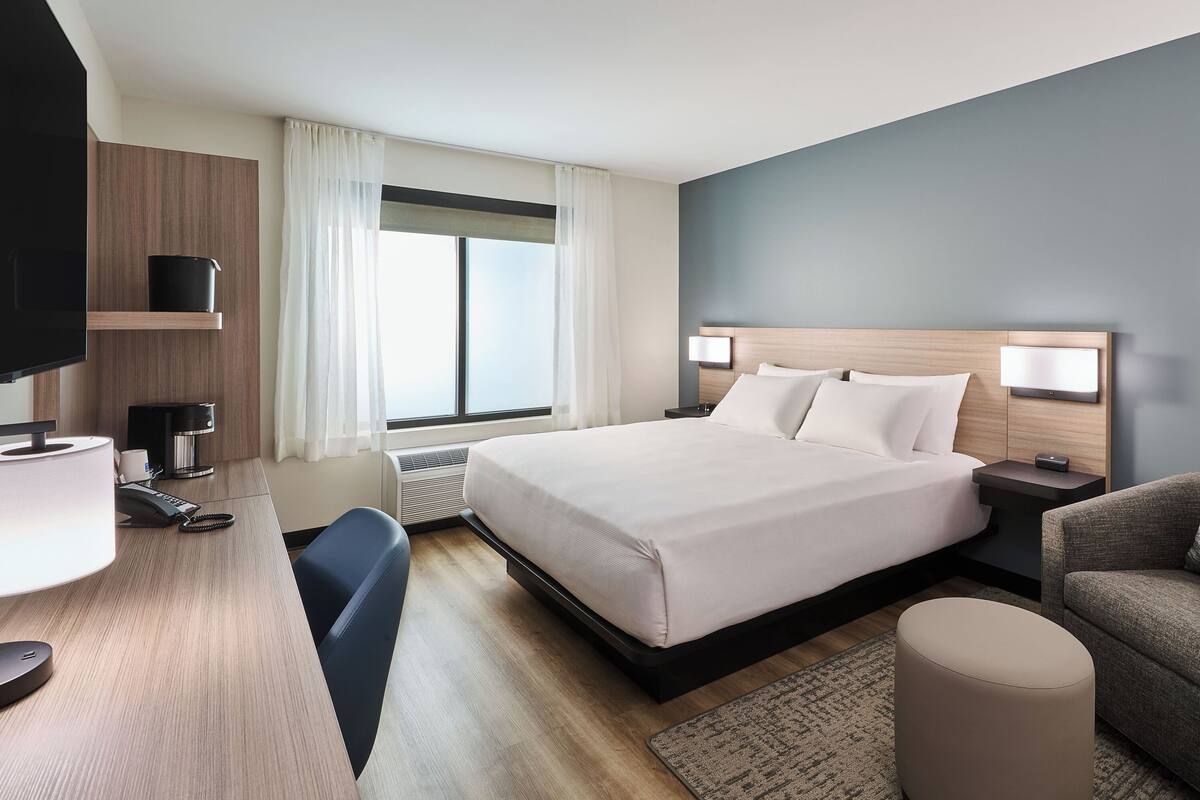 A hotel room with a bed, bedside tables, a desk with a lamp, a chair, a sofa, and a TV.