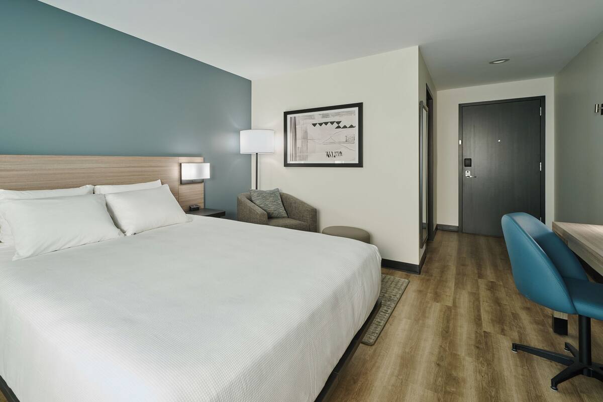 A hotel room with a bed, bedside table, lamp, chair, desk, and a framed picture on the wall.