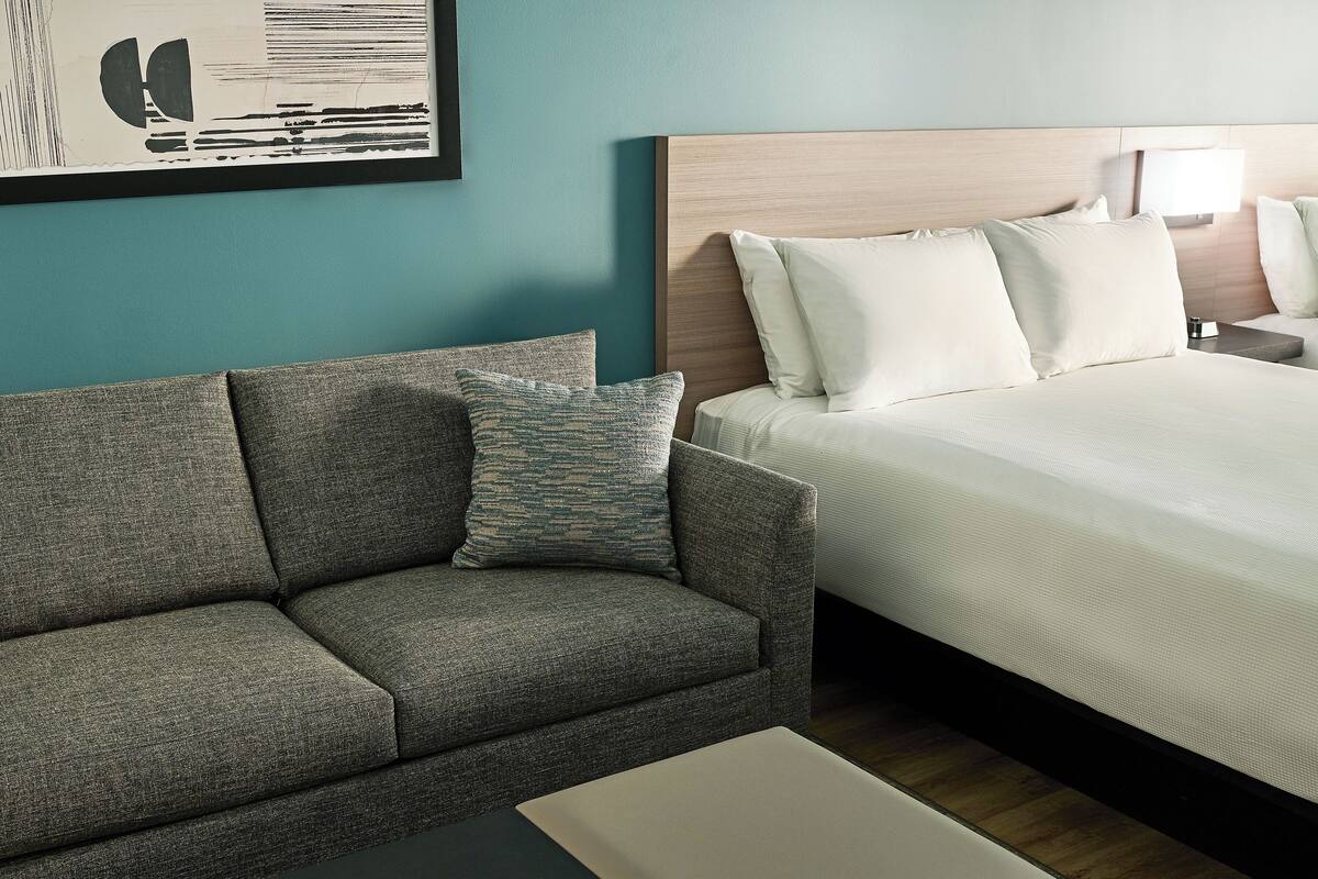 A hotel room with a bed, a sofa, a nightstand, a lamp, and a framed picture on the wall.