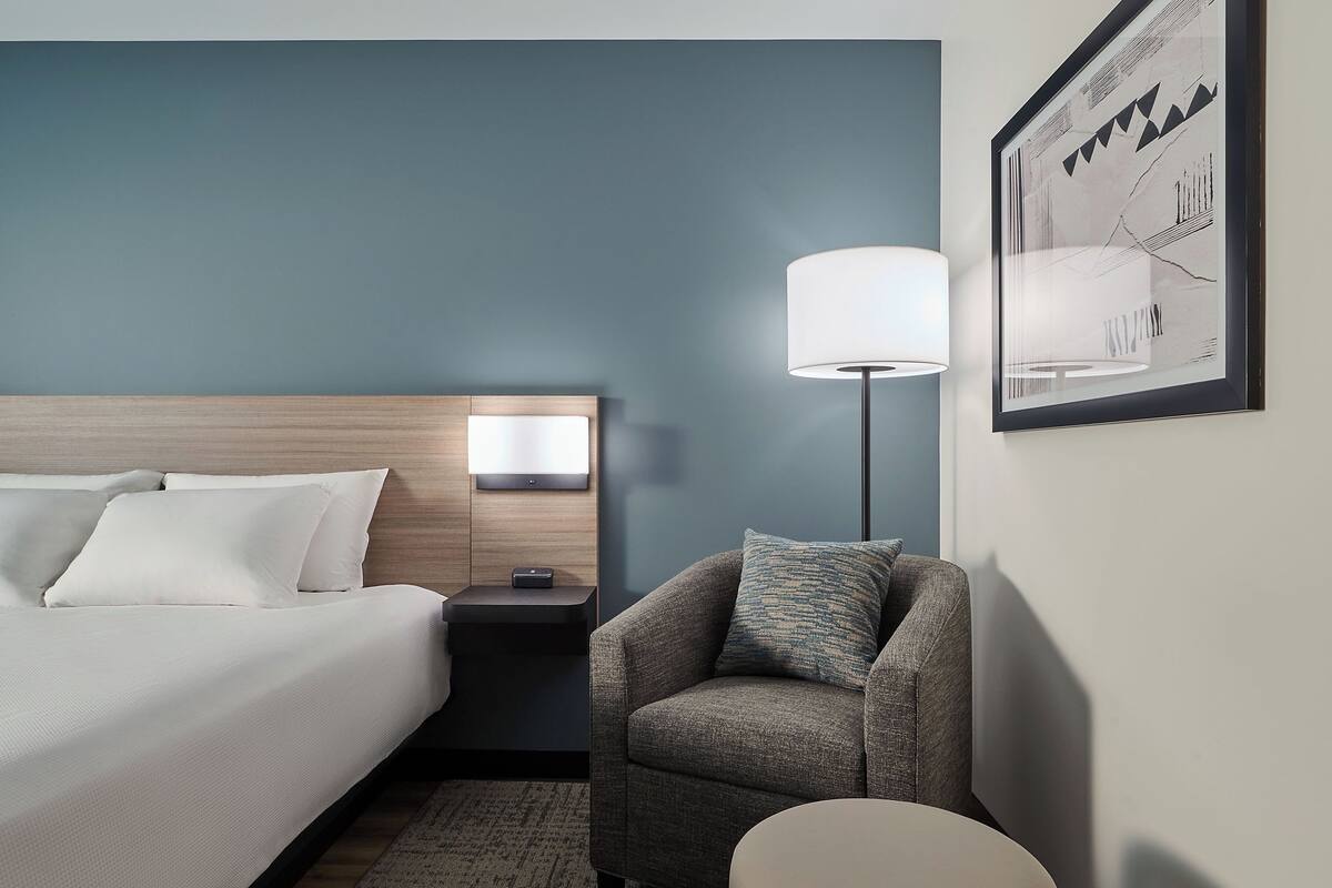 A hotel room with a bed, a nightstand, a lamp, a chair, and a rug.