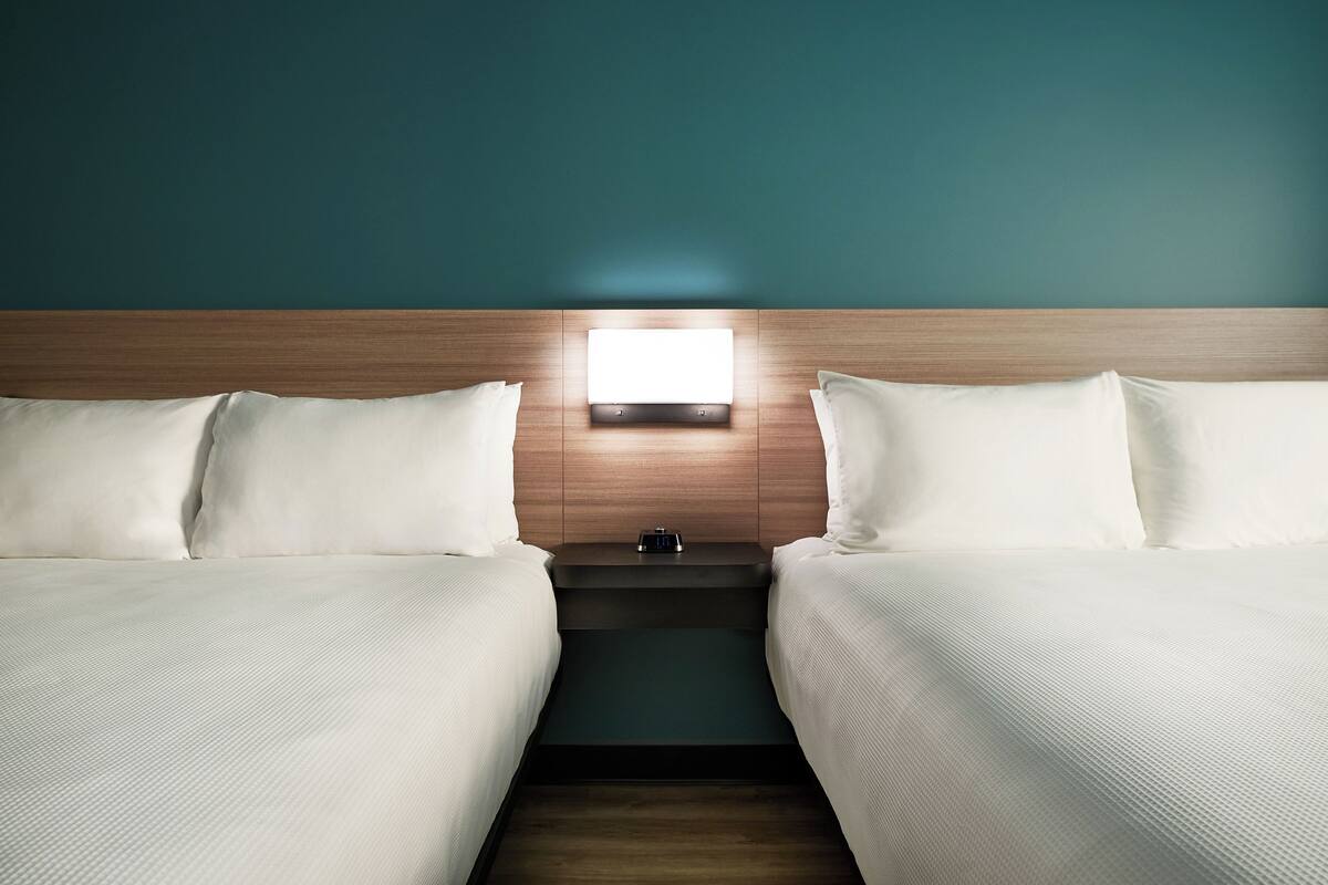 A hotel room with two beds, a nightstand, and a wall-mounted light.
