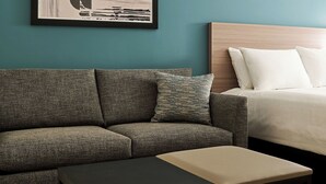 Una habitación de hotel moderna con un sofá gris, una mesita, una cama con ropa blanca y una obra de arte abstracta enmarcada en la pared.