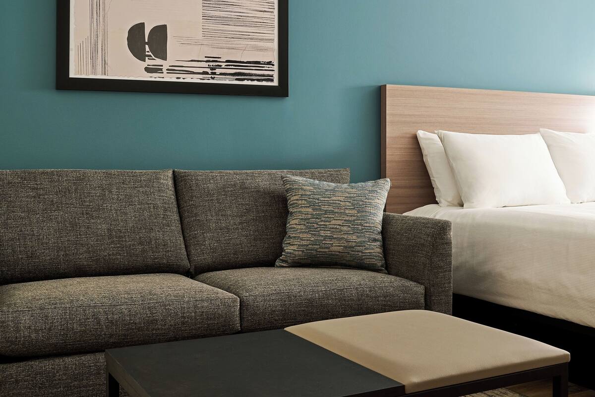 A modern hotel room with a gray sofa, a small table, a bed with white linens, and a framed abstract artwork on the wall.