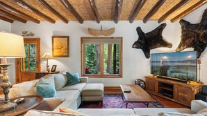 A cozy living room with a fireplace, a television, and a large window overlooking a forest. - Cabin on the River- Walk to Main St (Grand Lake)