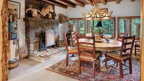 A rustic dining room with a stone fireplace, wooden ceiling beams, and a round wooden table with chairs. - Cabin on the River- Walk to Main St (Grand Lake)