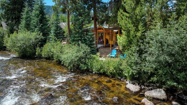 A cabin surrounded by dense forest, a flowing stream, and a clear blue sky. - Cabin on the River- Walk to Main St (Grand Lake)