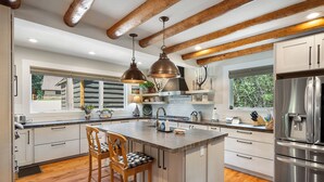 A modern kitchen with a central island, wooden beams, stainless steel appliances, and a view of a house outside. - Cabin on the River- Walk to Main St (Grand Lake)