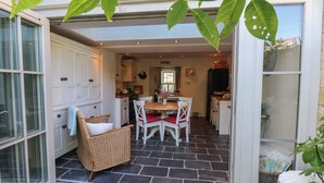 A dining area with a table and chairs, a wicker chair, and a view of the kitchen and living area through an open door. - Woodbine Cottage (Whitby)