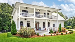 A two-story white house with a balcony on the upper floor, surrounded by a well-maintained lawn and trees. - Charming 3-bedroom farmhouse with Pool &Hot Tub in Asheville Fletcher area (Fletcher)