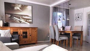 A modern living room with a sofa, a wooden cabinet, a dining table with chairs, and a large painting on the wall. - Vacation home Strandwind near Weißenhäuser Strand a.d. Baltic Sea with garden Oldenburg/H. (Oldenburg)
