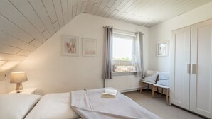 A bedroom with a sloped ceiling, a bed with a blanket, a bedside lamp, a chair, and a window with curtains. - Vacation home Strandwind near Weißenhäuser Strand a.d. Baltic Sea with garden Oldenburg/H. (Oldenburg)