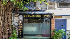 Ein Hotel namens "DE CLASSICO HOTEL" mit Glasfassade, blauer Tür und einem kleinen Gartenbereich mit Kunstgrashalm und Topfpflanzen.