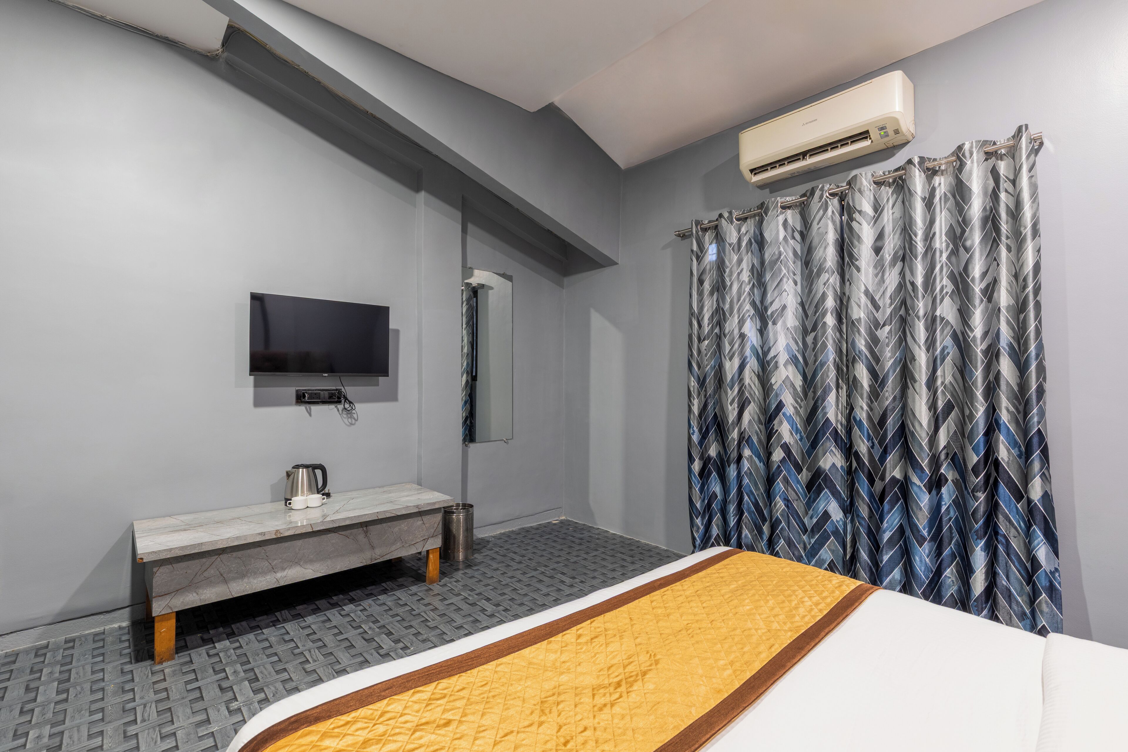 A hotel room with a bed, a bench, a television, an air conditioning unit, and a patterned curtain.