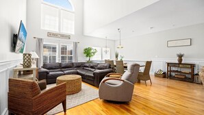 A spacious living room with a large L-shaped sectional sofa, a grey armchair, and a wicker ottoman. There is a dining area with wicker chairs and a wooden table. The room has large windows, a chandelier, and a sign that reads "SEASIDE HEIGHTS". - 3BR Seaside Haven - Beach Block Home w/ Parking (Seaside Heights)