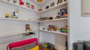 Une pièce aux murs blancs, avec un canapé rouge, une machine à coudre et des étagères remplies de livres et de jeux de société.