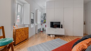 Une pièce moderne avec un plancher en bois, un téléviseur posé sur un meuble blanc, un canapé agrémenté de coussins à motifs et une cuisine en arrière-plan.