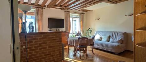 A cozy living room with a wooden ceiling, a dining area with a table and chairs, a sofa, and a TV mounted on the wall.