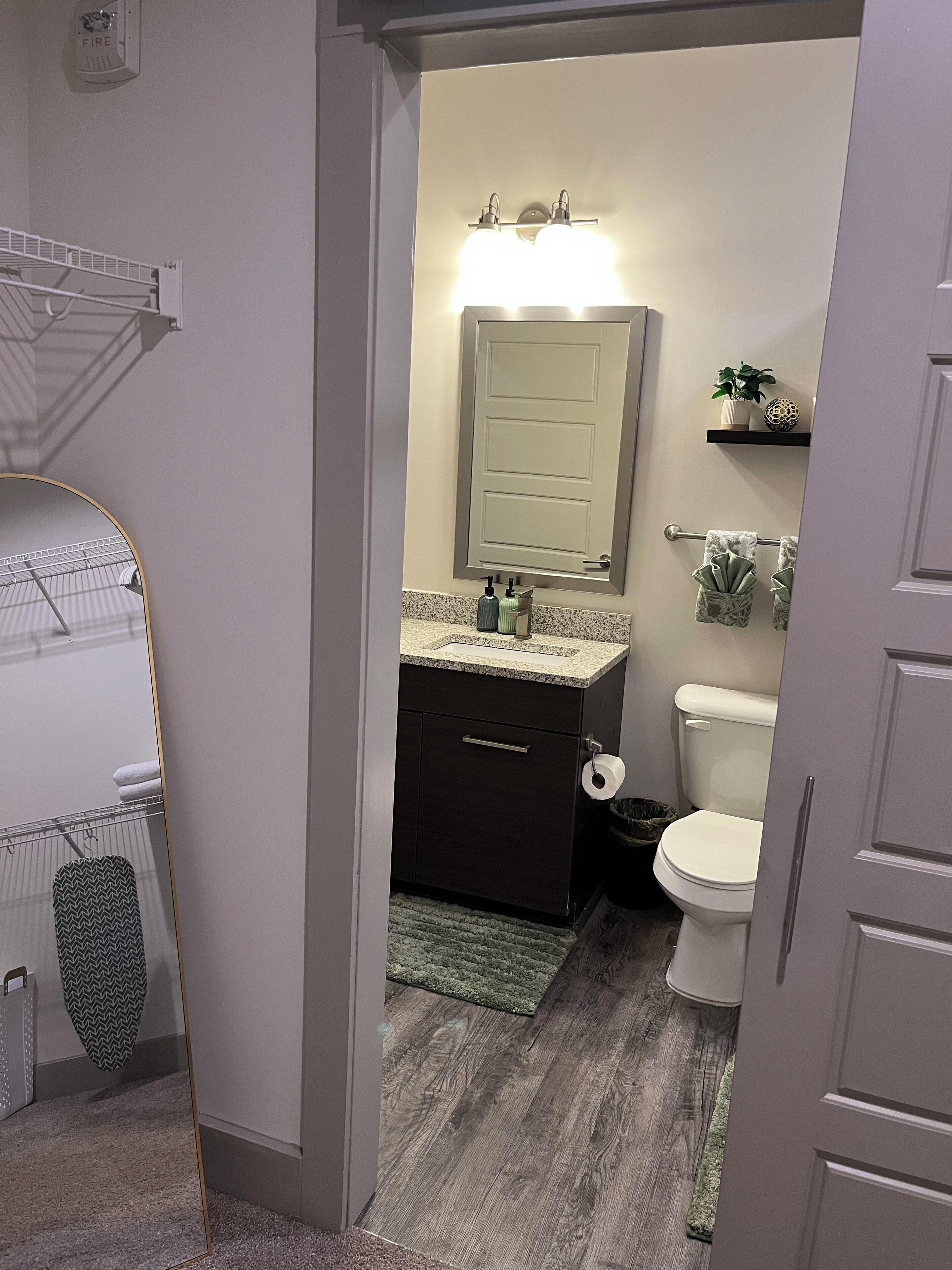 Une salle de bain comprenant des toilettes, un lavabo et un miroir. On y trouve un porte-serviettes avec des serviettes, une poubelle et une petite étagère avec des objets décoratifs.