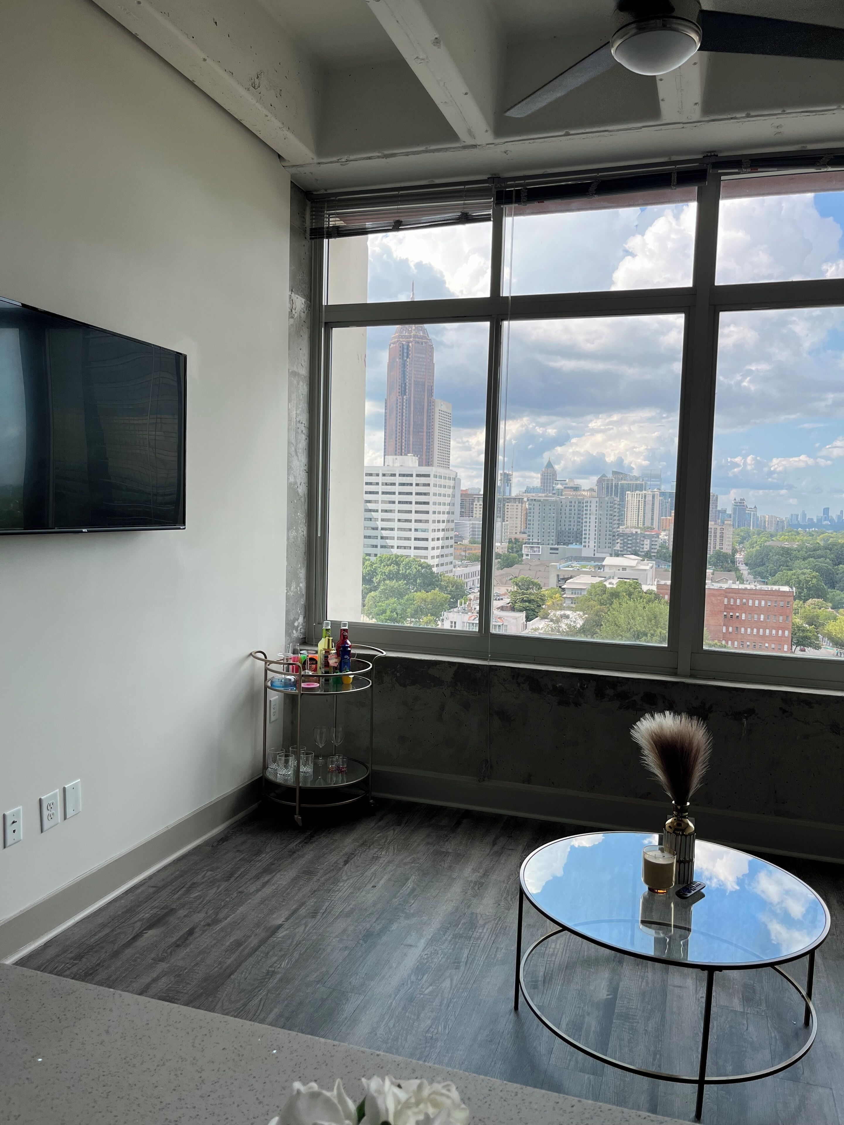 A room with a large window, a mounted TV, a small table with a vase, and a metal shelf with various items.