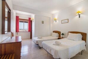 Habitación de hotel con dos camas, un escritorio de madera, una ventana con cortinas rojas y lámparas fijadas a la pared.