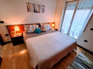A hotel room with a large bed, floral-patterned pillows, a nightstand with a lamp, and a view through a sliding glass door.