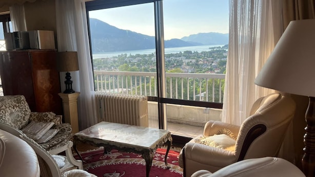 A spacious living room with a large window offering a scenic view of a lake and mountains. - Large "classic" apartment, breathtaking view. Tennis court and swimming pool nearby. (Tresserve)