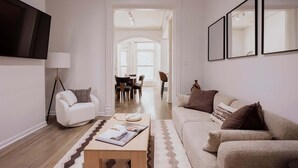 Sala de estar con sofá, mesa de centro, lámpara y televisor de pantalla plana. Se observa un comedor al fondo, con una mesa y sillas.