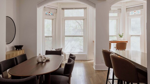 Un comedor con una mesa y sillas de madera, paredes blancas y ventanas grandes con marcos blancos.