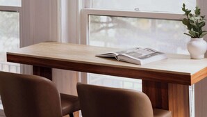 Una mesa de madera con dos sillas, un libro y una planta en maceta cerca de una ventana.