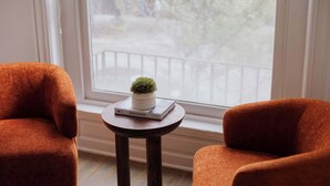 Une pièce avec deux fauteuils orange, une petite table en bois sur laquelle se trouvent un livre et une plante en pot, et une fenêtre donnant sur un balcon.