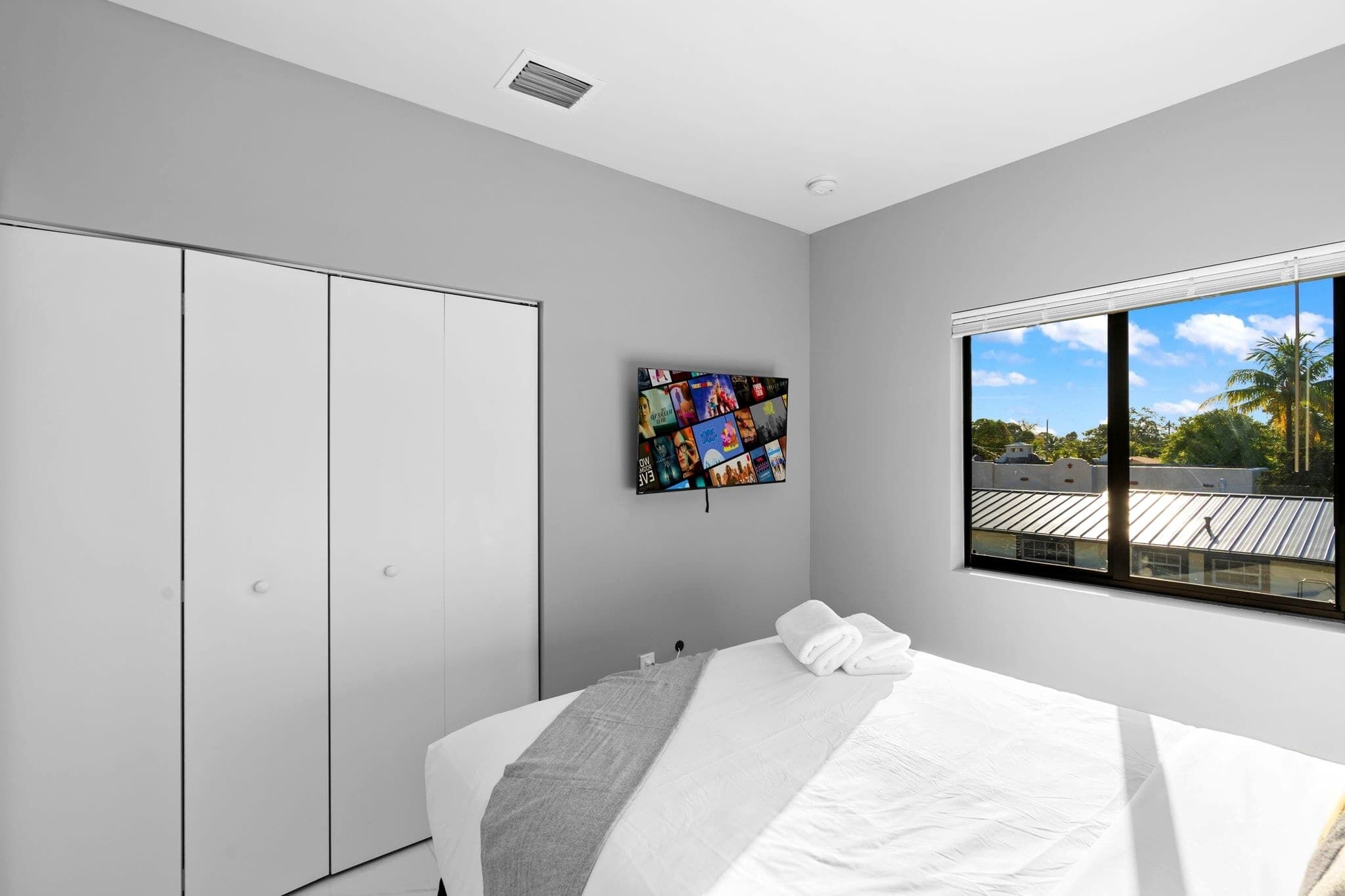 A modern bedroom with a large bed, a wardrobe, a wall-mounted TV, and a window with a view of the sky and palm trees.