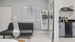 Un salon moderne avec un canapé gris, une porte blanche, une fenêtre avec des stores et un coin cuisine équipé d’un four à micro-ondes et d’une machine à café.