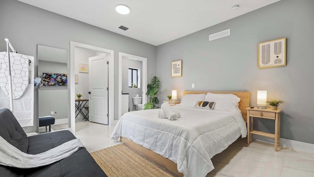 Une chambre moderne avec un grand lit, une table de chevet avec une lampe, une chaise et une vue sur la salle de bain et la cuisine.