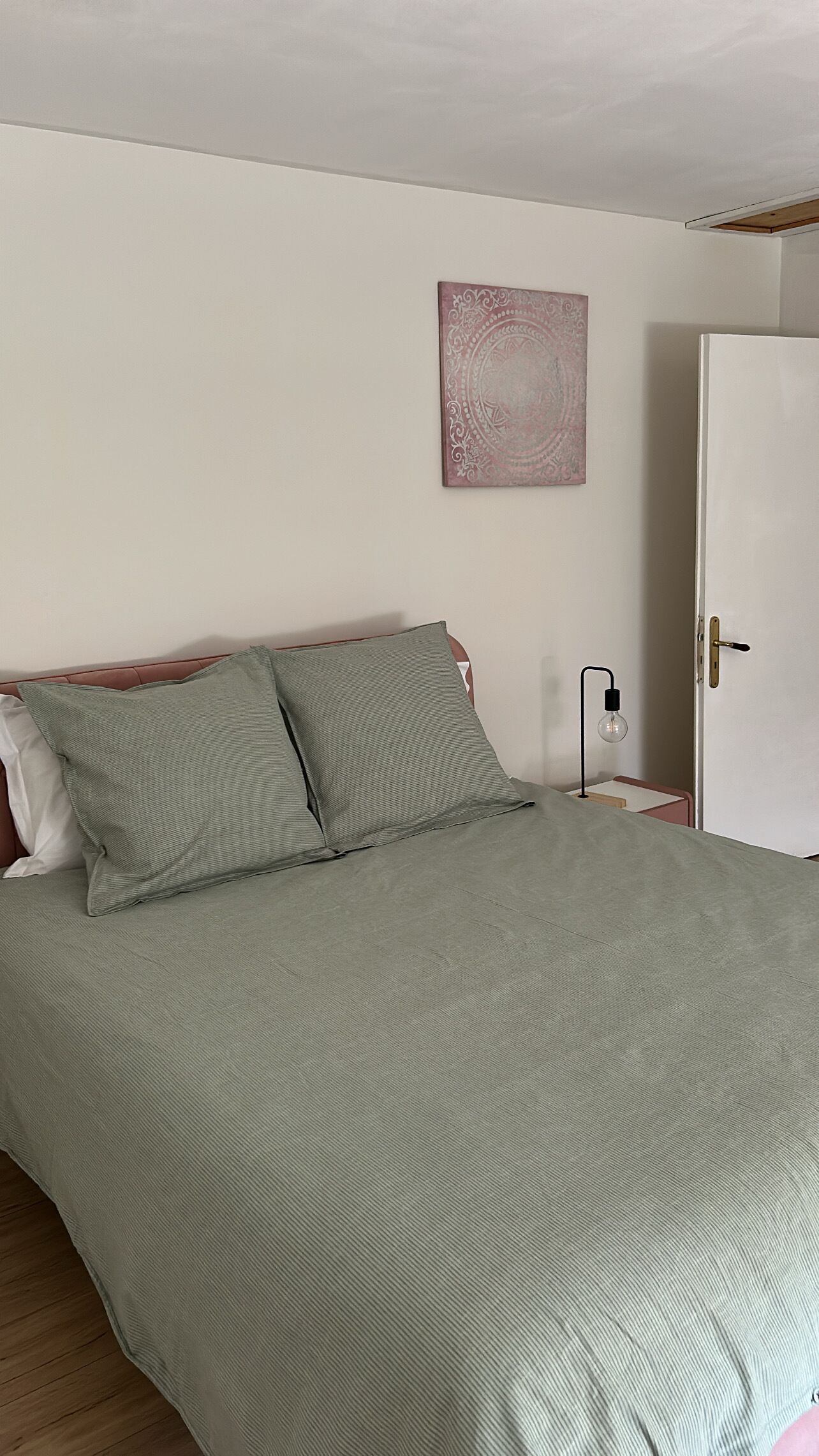 Een bed met een groene sprei, twee kussens, een kunstwerk aan de muur, een bedlampje en een deur.