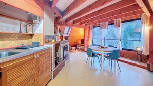 A cozy cabin interior with a wooden dining table and chairs, a kitchen area with a sink and cabinets, and a large window providing a view of the outdoors. - Chalet BomBom - Cozy cocoon with pond view in Mittlach (Mittlach)