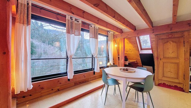 A wooden interior with a round table and chairs, large windows with curtains, and a view of trees outside. - Chalet BomBom - Cozy cocoon with pond view in Mittlach (Mittlach)