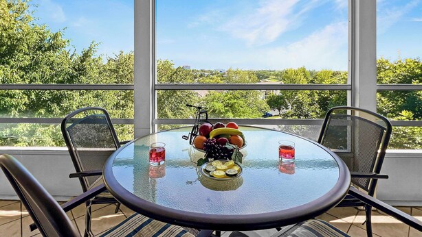 A round glass table with chairs, a fruit bowl, and two glasses of red drink on a balcony with a view of greenery and buildings.