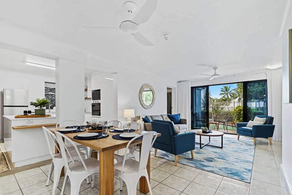 A modern living room with a dining area, featuring a large window, a ceiling fan, and a view of the outdoors.