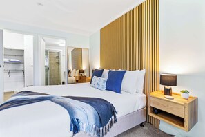 A modern bedroom with a large bed, a wooden headboard, a bedside table with a lamp, and a mirror. - Oceanfront 3 Bedroom Apartment with Pool in Cairns (Cairns North)