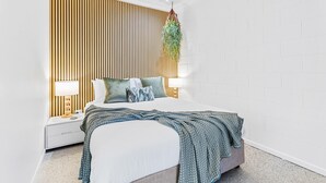 A modern bedroom with a wooden slat wall, a bed with a blue blanket, two bedside tables with lamps, and a hanging plant.